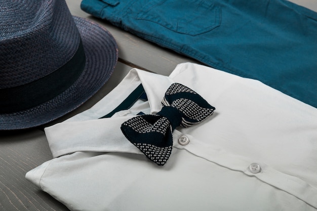 Roupa masculina com fundo de madeira, roupas da moda infantil, fedora cinza, calça azul marinho, camisa branca, gravata borboleta preta, vista superior, postura plana, espaço de cópia.