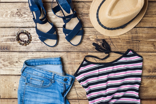 Roupas femininas de verão. foto de moda plana leigos. blue jeans, t-shirt, chapéu de sol, sandálias azuis sobre fundo de madeira.