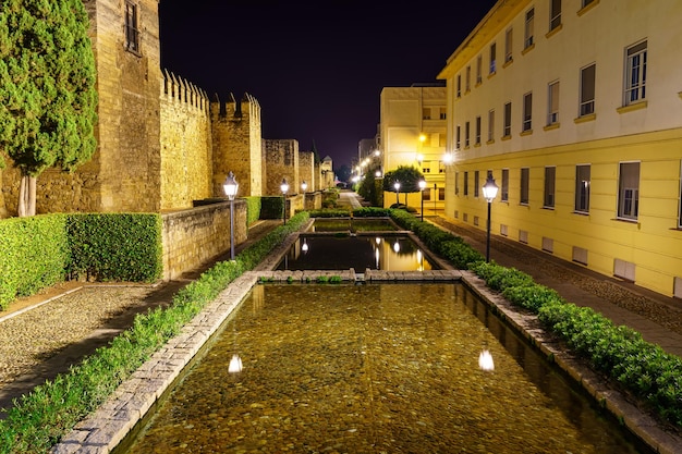 Rua iluminada à noite ao longo da parede com jardins e fontes. Córdoba, Espanha.