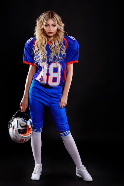 Rubia atlética haciéndose pasar por chica de fútbol americano sobre fondo negro. Mujer joven hermosa que lleva la bola que se sostiene superior del fútbol americano. Foto de estudio de mujer de fútbol americano