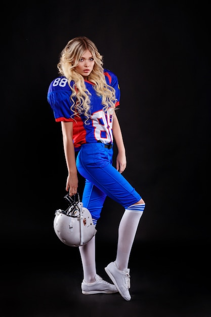 Rubia atlética haciéndose pasar por chica de fútbol americano sobre fondo negro. Mujer joven hermosa que lleva la bola que se sostiene superior del fútbol americano. Foto de estudio de mujer de fútbol americano