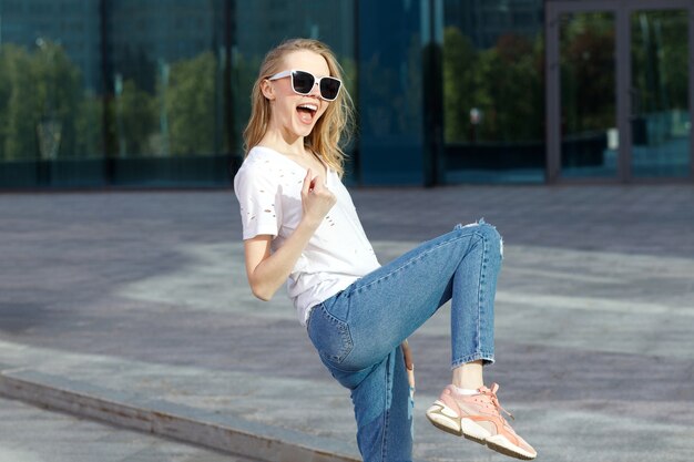 Rubia atractiva emocional con gafas y camiseta blanca muestra emociones sobre un fondo azul en el verano