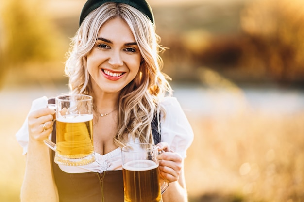 Rubia bastante feliz en vestidos típicos, vestido de fiesta tradicional, sosteniendo dos jarras de cerveza al aire libre en el campo