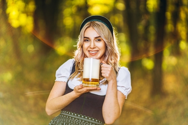 Foto rubia bastante feliz en vestidos típicos, vestidos de fiesta tradicionales, sosteniendo una jarra de cerveza al aire libre en el bosque