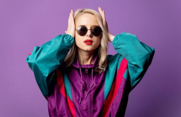 Rubia elegante en cazadora de los años 80 y gafas de sol redondas en la pared púrpura