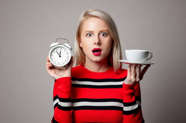 Rubia con estilo en suéter rojo con reloj despertador y taza de café en la pared gris