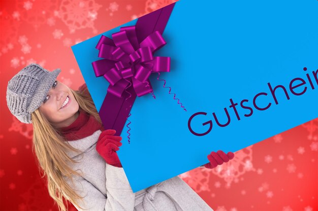 Rubia feliz con ropa de invierno que muestra la tarjeta contra el delicado diseño de copos de nieve generado digitalmente