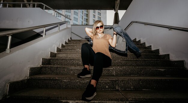 La rubia con gafas de sol lleva una chaqueta de vaqueros sentada en las escaleras chica moderna