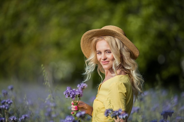 Rubia joven hermosa mujer con un sombrero camina por un campo de flores de color púrpura. El verano. Primavera.