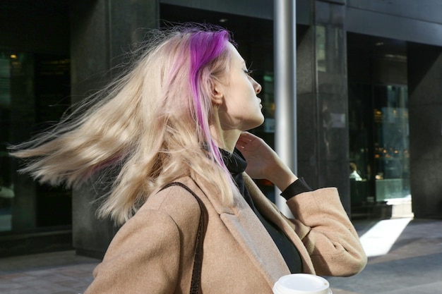 rubia con mechones de cabello lila en un abrigo beige con una taza de café en la mano en th