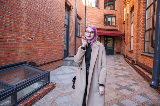 rubia con mechones de pelo lila en un abrigo beige y gafas en el fondo de una construcción de ladrillo rojo