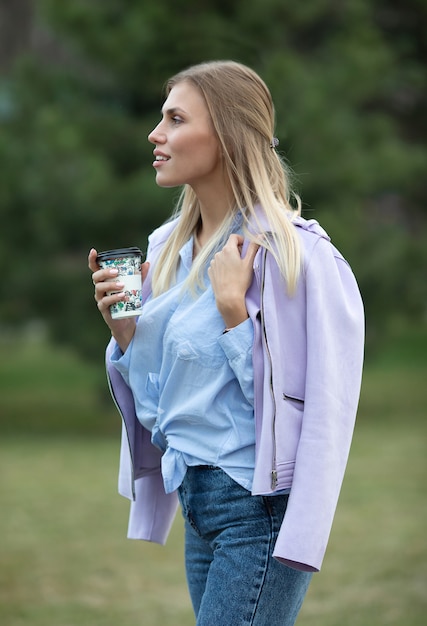 Foto rubia de moda alegre que sostiene el café al aire libre. mujer joven que bebe el café en la calle de la ciudad.