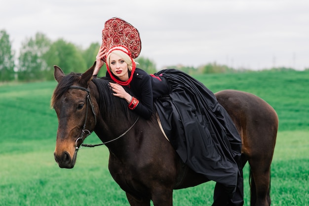 Rubia mujer eslava en vestido negro y tocado kokoshnik en campo con un caballo negro al atardecer