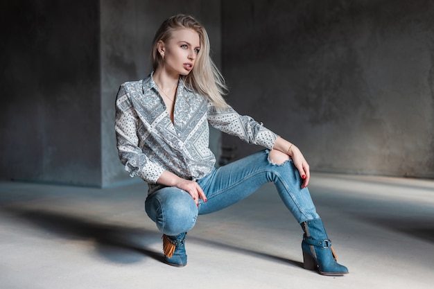 Rubia de mujer joven moderna de moda en camisa elegante en jeans rotos de moda en botas de vaquero vintage posando sentado en una habitación con luz solar