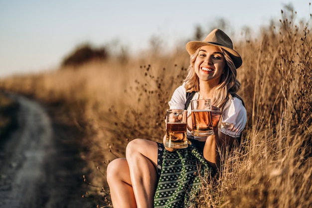 Rubia muy feliz en vestidos típicos, vestidos de fiesta tradicionales, sentada con dos jarras de cerveza al aire libre en el campo