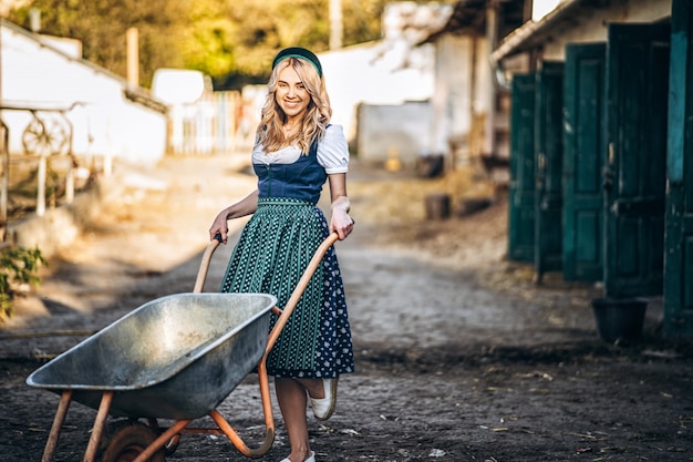 Rubia muy sonriente en traje tradicional con carretilla trabajando en el patio trasero.