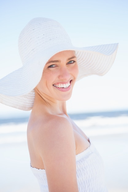 Rubia muy sonriente en vestido blanco y sombrero para el sol en la playa