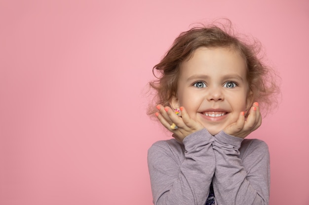 Rubia de niña pequeña con niña rizada se pintó los dedos con esmalte de uñas, mira en el marco en el estudio y sonríe.