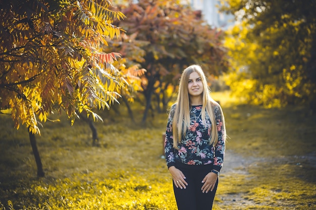 Rubia de pelo largo de niña mujer en un callejón con árboles con hojas rojas. Ella está feliz confiada en una sesión de fotos. Parque de otoño soleado día cálido.