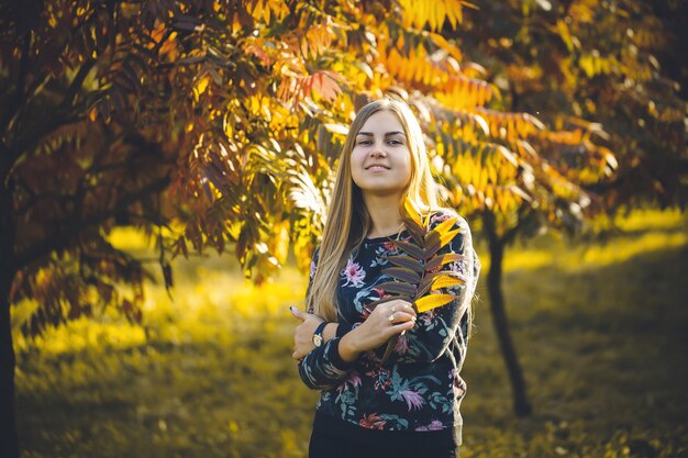 Rubia de pelo largo de niña mujer en un callejón con árboles con hojas rojas. Ella está feliz confiada en una sesión de fotos. Parque de otoño soleado día cálido.