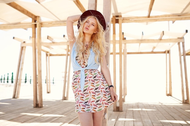 Foto rubia con el pelo rizado vestida con un vestido ligero de verano y un sombrero posando en la terraza de madera mirando a la cámara