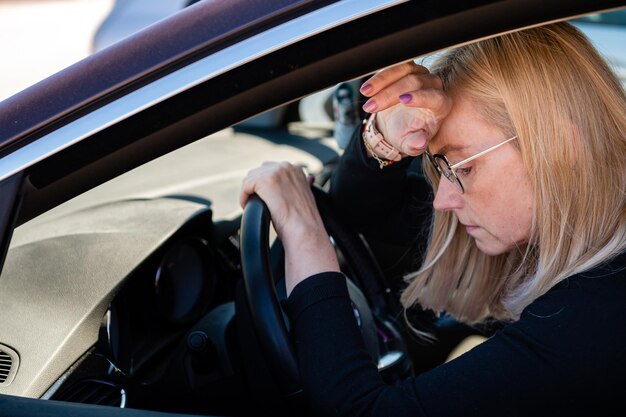 Una rubia pensativa de mediana edad con gafas se sienta al volante de un auto