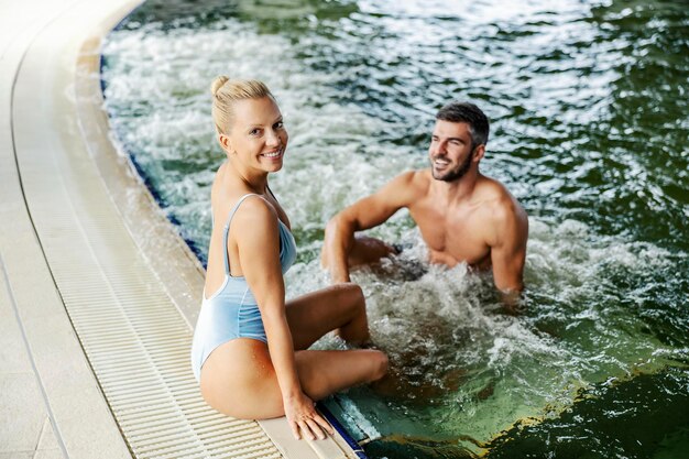 Una rubia sexy está sentada junto a una piscina con agua termal y mirando a la cámara.