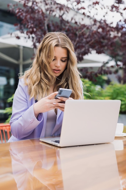 Foto rubia sexy con un traje elegante escribe un mensaje en su teléfono mientras trabaja afuera desayuno en la terraza de verano