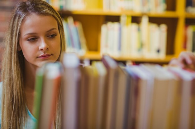 Rubia sonriente escogiendo un libro de estanterías en la biblioteca