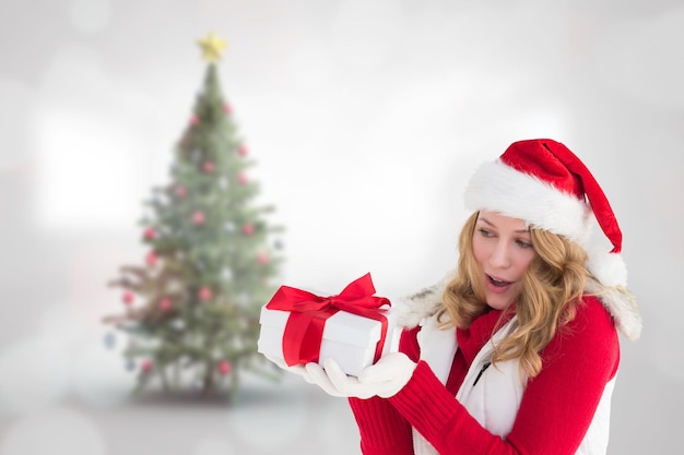 Rubia sorprendida con sombrero de santa con regalo contra el árbol de navidad borroso en la habitación