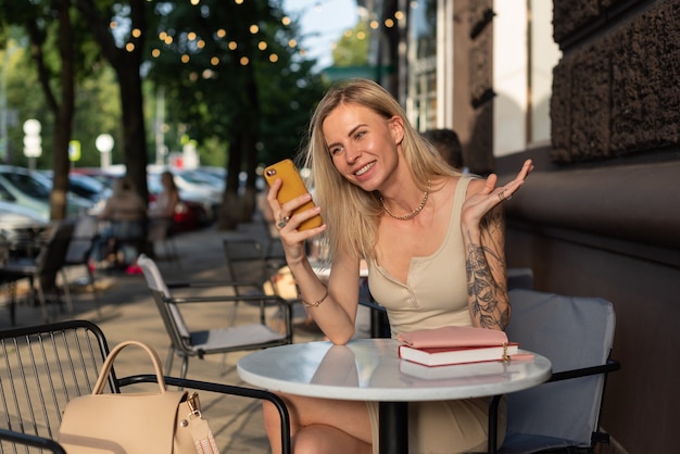 Una rubia con un tatuaje en el brazo se sienta en un café de verano y habla emocionalmente por teléfono.
