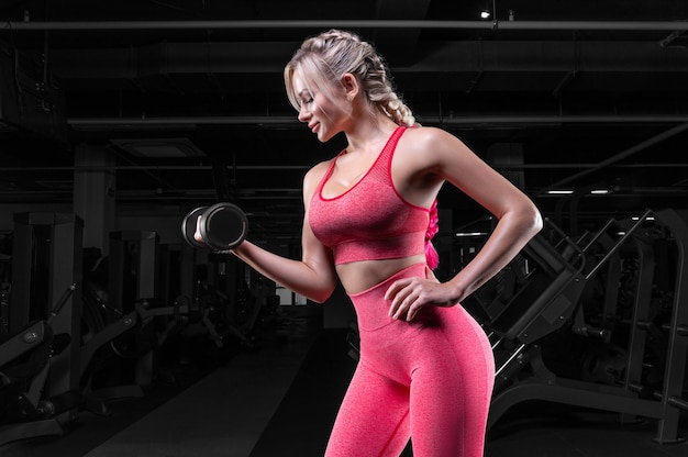 Rubia tetona en chándal rosa posando en el gimnasio con mancuernas. Concepto de fitness.