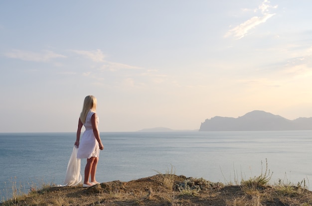 Rubia con un vestido blanco de pie en la montaña y mira al mar