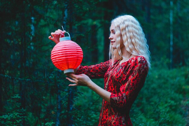 Una rubia con un vestido rojo en el bosque con una linterna china roja en las manos