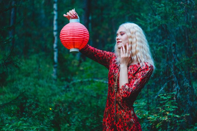 Una rubia con un vestido rojo en el bosque con una linterna china roja en las manos