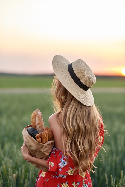 Una rubia con un vestido rojo y un sombrero de paja con pan francés en sus manos en un campo de trigo al atardecer