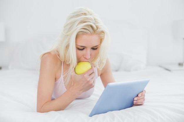 Rubio informal comiendo una manzana mientras usa la tableta en la cama