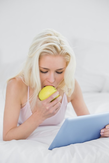Rubio informal comiendo una manzana mientras usa la tableta en la cama