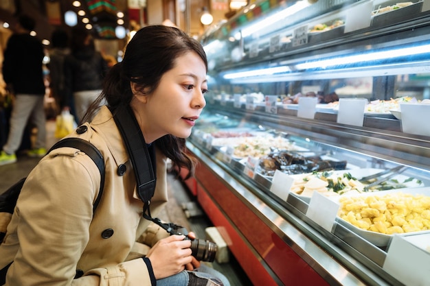 Rucksacktouristin mit Kamera betrachtet eine Vielzahl von Feinschmeckern im Feinkostbereich in Japan. Chinesisches Mädchen mit Digitalkamera wählt aus einer Sammlung von Lebensmitteln aus, die in einer Vitrine ausgestellt sind.