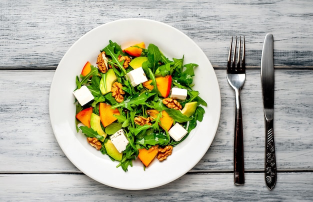 Rucola-Salat, Persimone, Walnüsse, Avocados, Feta-Käse auf einem weißen Teller auf einem Holztisch