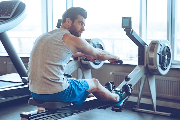 Rudern im Fitnessstudio. Seitenansicht eines jungen Mannes in Sportkleidung, der vor dem Fenster im Fitnessstudio rudert und wegschaut