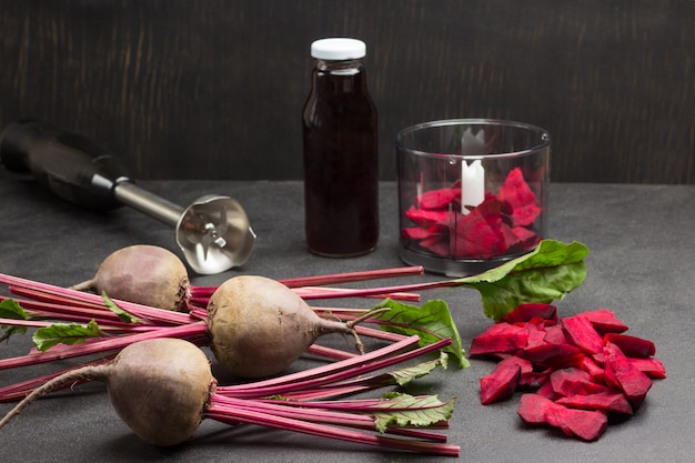 Rüben mit Spitzen, geschnittene Rüben. Rote-Bete-Saftflasche. Geschnittene Rüben im Mixglas. Cutter. Draufsicht. Schwarzer Hintergrund