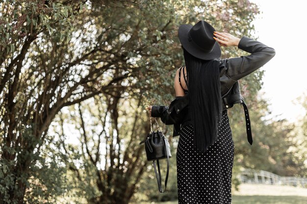 Rückansicht auf modische Frau mit gesundem, langem, schickem Haar in elegantem schwarzem Hut in schönem Kleid in Vintage-Jacke mit Lederhandtasche im Park. Trendiges Mädchen in modischer schwarzer Frühlingskleidung auf dem Weg.