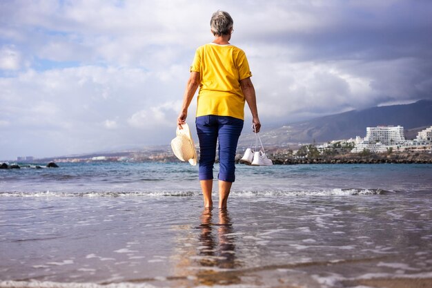 Rückansicht der älteren Frau barfuß am Meer, die Schuhe und Hut in der Hand hält, während sie am schwarzen Sandstrand steht Ältere Frau, die Urlaub und Freiheit genießt