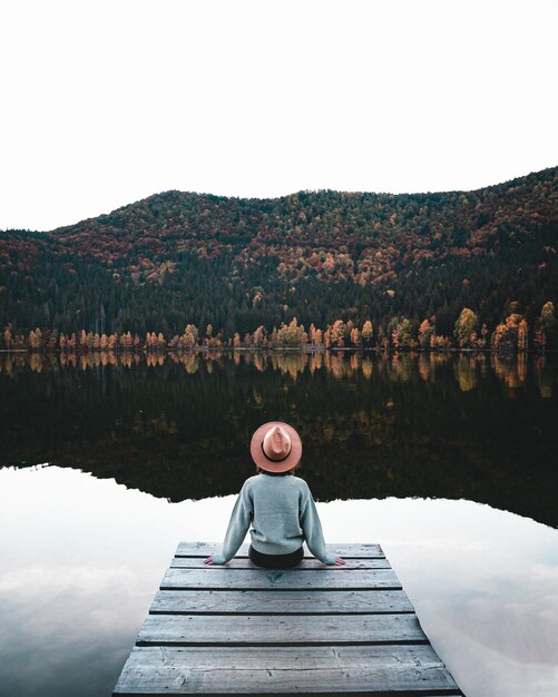 Foto rückansicht der altmodischen jungen frau, die auf einem holzsteg sitzt und seeblick ansieht weiblicher hipster mit braunem hut entspannt sich am rande des stegs und bewundert die herbstfarben