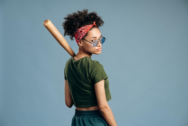 Rückansicht der ernsthaften brutalen Frau, die mit Baseballschläger posiert und mit ruhigem Ausdruck in die Kamera schaut. Sportgerätekonzept