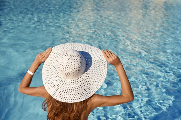 Rückansicht der Frau im weißen Hut, der nahe Pool an einem sonnigen Tag sitzt. Seereisekonzept