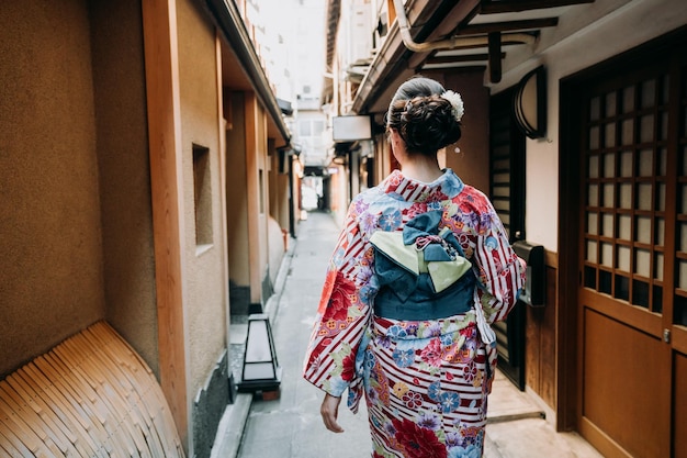 Rückansicht der jungen Dame in traditioneller japanischer Kleidung auf der Straße, die an der hölzernen Tür des Papierfensters in der historischen Altstadt von Kyoto, Japan, vorbeigeht. schönes Mädchen in buntem Blumenkimonostoff.