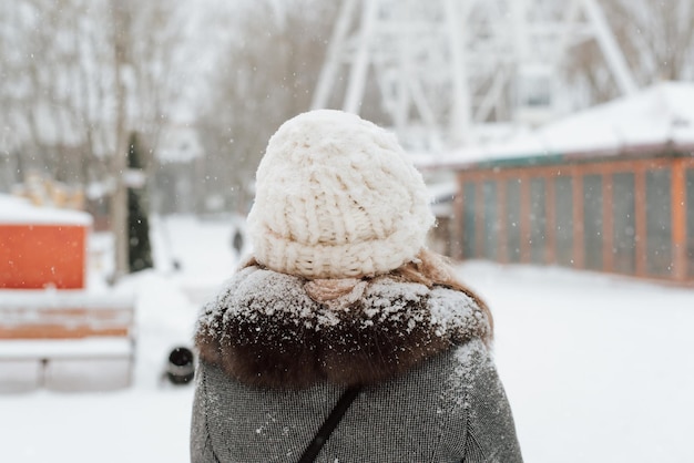 Rückansicht der jungen Frau, die im Winter Schneefälle, verschneites Wetter im Park genießt. Rückansicht des Mädchens in Mantel und gemütlicher weißer Strickmütze, die im Freien steht