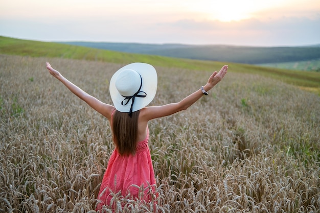Rückansicht der jungen glücklichen Frau in rotem Sommerkleid und Strohhut, die auf gelber Bauernhofwiese steht, mit reifem goldenem Weizen, der ihre Hände anhebt und einen warmen Abend genießt.
